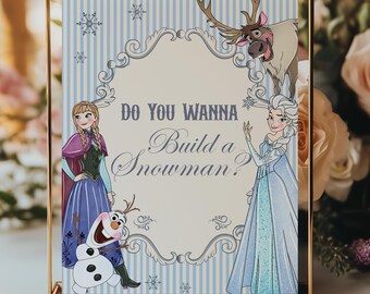 May include: A framed sign with the text "Do You Wanna Build a Snowman?" features illustrations of characters from the movie Frozen. The sign has a light blue and white striped background and is in a gold frame.