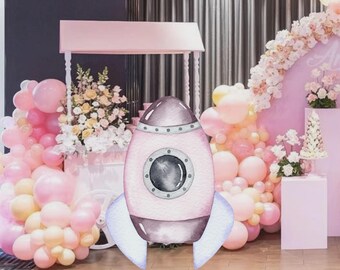 Puede incluir: Un cohete rosa y gris en acuarela con una ventana redonda y aletas azul claro. El cohete está frente a una escena de fiesta con globos rosas y amarillos, flores y un carrito rosa.