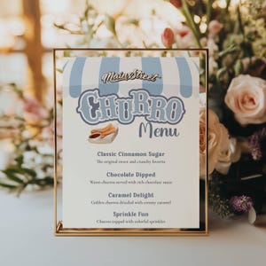 May include: A gold-framed menu for MainStreet Churro, featuring a light blue awning design. The menu lists churro flavors: Classic Cinnamon Sugar, Chocolate Dipped, Caramel Delight, and Sprinkle Fun. The menu is displayed in front of a floral arrangement.