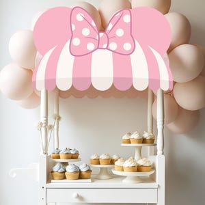 May include: A white cupcake cart with a pink and white striped awning, topped with a pink bow with white polka dots. The cart is decorated with cupcakes and balloons, creating a festive atmosphere.
