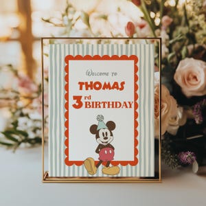 Puede incluir: Un letrero de cumplea&ntilde;os enmarcado con un borde dorado. El letrero tiene un borde rojo festoneado y un fondo a rayas azul claro y blanco. El texto dice "Bienvenido al 3er cumplea&ntilde;os de Thomas" con un gr&aacute;fico de Mickey Mouse.