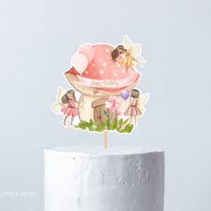 Op de afbeelding: Een roze en witte paddenstoel taarttopper met drie feeën en de tekst "Happy Birthday".