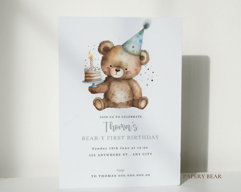 May include: A birthday invitation featuring a watercolor illustration of a brown teddy bear wearing a party hat and holding a birthday cake with a lit candle. The text on the invitation reads "Join us to celebrate Thoma's Bear-Y First Birthday, Sunday 18th June at 12:30, 123 Anywhere St., Any City, rsvp to Thomas 000.000.000.00, Papery Bear".