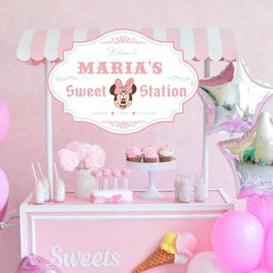 May include: A pink and white candy cart with a sign that says "Welcome to Maria's Sweet Station". The sign features a cartoon image of Minnie Mouse. The cart is decorated with pink and white balloons, cupcakes, and lollipops.