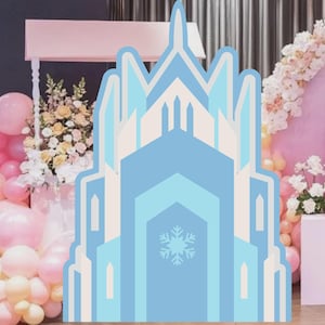 Peut inclure: Décoration de fête en forme de château bleu clair avec un motif de flocon de neige. La toile de fond comprend des ballons roses et jaunes, des compositions florales et une arche rose, créant une ambiance festive.