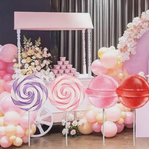 May include: A whimsical scene featuring a pink candy cart, balloons, and floral arrangements. Four large lollipops in purple, pink, and red are on display. The lollipops are on sticks, adding a playful touch to the party decor.