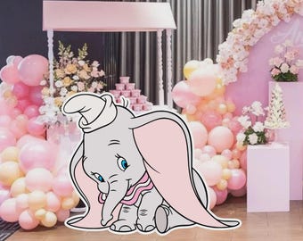 May include: A cutout of Dumbo the elephant, in gray with pink ears and a pink collar, sits in front of a pink and white party backdrop. The backdrop includes balloons, flowers, and a dessert table, creating a festive atmosphere.