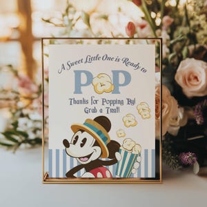 Könnte beinhalten: Ein gerahmtes Schild mit dem Text "A Sweet Little One is Ready to POP. Thanks for Popping By! Grab a Treat!" zeigt eine Cartoon-Figur von Mickey Mouse, die Popcorn hält. Das Schild hat einen goldenen Rahmen und einen blau-weiß gestreiften Hintergrund.