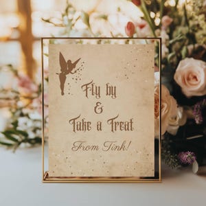 May include: A framed sign with a gold border and a vintage-style background. The sign features a silhouette of a fairy and the text "Fly by & Take a Treat From Tink!" in brown script. Decorative stars are scattered across the sign.