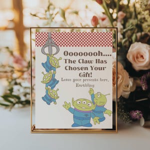May include: A gold framed sign with the text "Oooooooh.... The Claw Has Chosen Your Gift! Leave your presents here, Earthling." The sign features cartoon aliens and a claw graphic, with a red and white checkered border.