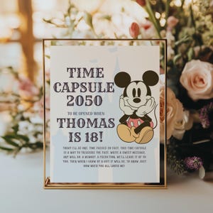 May include: A gold framed sign with the text "TIME CAPSULE 2050" and "THOMAS IS 18!" featuring a Mickey Mouse illustration. The sign is a keepsake for a future birthday.