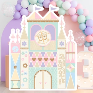 May include: A colorful, whimsical castle-shaped party decoration with pastel colors, including pink, blue, and yellow. The design features a clock face, Mickey Mouse silhouettes, and the numbers 2, 5, 8, 7, 4, 6, and 3. A balloon arch and a light-up number 3 are in the background.