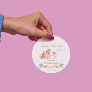 Może przedstawiać: Okrągła, biała naklejka z napisem "Bibbidi Bobbidi Boo!" oraz grafiką myszy, dyni i banera. Naklejka jest trzymana na różowym tle.