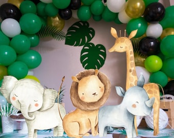 Op de afbeelding: Een jungle-themafeest met een boog van groene, gouden, zwarte en witte ballonnen. Waterverf dierenuitsparingen van een olifant, leeuw, neushoorn en giraffe zijn te zien. Groene monstera bladeren dragen bij aan de decoratie.