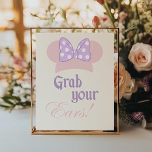 Può includere: Un cartello con cornice dorata presenta un design a tema Minnie Mouse. Il design include una silhouette rosa della testa di Minnie, un fiocco viola con pois bianchi e il testo "Grab your Ears!" in viola e rosa.