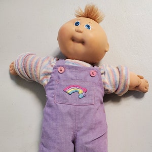 May include: A Cabbage Patch Kid doll with blonde hair and blue eyes wearing a purple overall with a rainbow patch and a striped shirt.