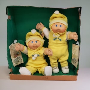 May include: Two Cabbage Patch Kids dolls wearing yellow winter outfits with white hats and white boots. The dolls are sitting in a green and brown box with the words "Cabbage Patch Kids Twins" printed on the side.