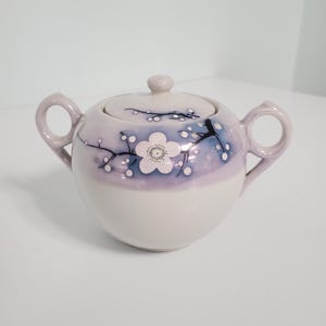 May include: A white ceramic sugar bowl with a lid, featuring a floral design in shades of blue and purple. The design includes a large white flower with a black center and smaller white flowers on branches.