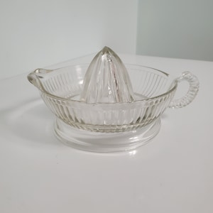 May include: A clear glass citrus juicer with a ribbed bowl and a pointed reamer. The juicer has a handle on each side.