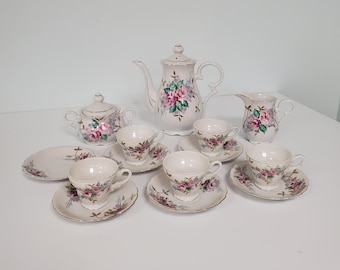May include: A vintage porcelain tea set featuring a teapot, creamer, sugar bowl, and several teacups with saucers. The set is decorated with pink and purple roses and gold trim, creating an elegant aesthetic.