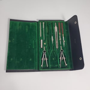 May include: A black leather case with a green felt interior holds a set of drafting tools, including compasses, rulers, and a pencil.