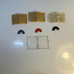 May include: An assortment of vintage bird calls and accessories. Three small, folded paper pamphlets with the text "Swiss Warbler Bird Call" are arranged above three semi-circular, fan-shaped pieces in black, red, and black. A clear plastic case is at the bottom.