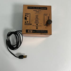 May include: A vintage model train power pack, labeled "FOR ALL HO TRAINS." The orange metal box has a dial for speed control and switches for reverse and forward. It includes a black power cord and plug.