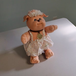 May include: A pink plush dog with a white fluffy head, wearing a white dress with pink hearts and a brown collar with a gold charm.