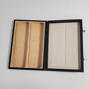 May include: Black wooden box with a hinged lid, containing rows of slots for storing slides. The lid has a numbered grid for organizing the slides.