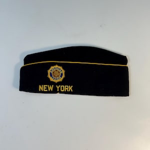 May include: Black military-style cap with gold trim and embroidered text. The cap features a circular emblem with the words "American Legion" and the state name "New York" in gold lettering. The cap is likely a commemorative item.