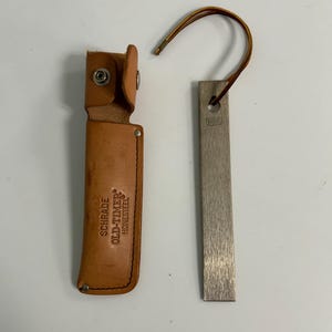 May include: A leather sheath with a snap closure and the text "SCHRADE OLD-TIMER HONESTEEL". Next to it is a metal sharpening tool with a leather loop. The sharpening tool is rectangular and has the text "SCHRADE" printed on it.