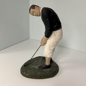 May include: A vintage figurine of a golfer in a classic pose, dressed in a black jacket, white pants, and black shoes. The figure is bent over, focused on the golf club and ball on a green, textured base. The figurine is likely made of a composite material.