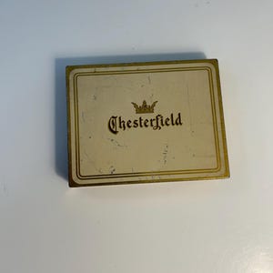 May include: A rectangular, vintage Chesterfield tin with a cream-colored background and gold trim. The word "Chesterfield" is printed in a dark brown, stylized font, topped with a gold crown emblem. The tin shows signs of wear.