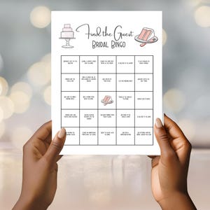 May include: A white bridal bingo game card held by hands. The card features a cake illustration and the text "Find the Guest BRIDAL BINGO". The bingo grid includes various wedding-related prompts, such as "Shares a favorite memory with the bride."