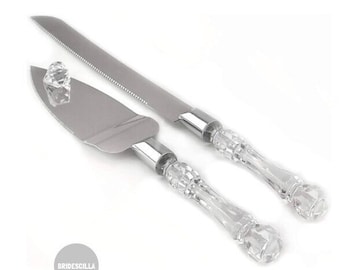 May include: A set of two silver cake cutting knives with clear plastic handles. The handles have a decorative, ornate design.