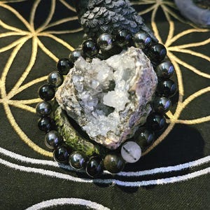 Peut inclure: Une figurine de corbeau gris foncé aux yeux rouges est posée sur une géode et un bracelet de perles. Le bracelet comporte des perles noires et argentées, et un cristal blanc et transparent. Le corbeau est sur une surface noire et dorée.