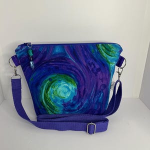 May include: A crossbody bag featuring a swirling abstract pattern in shades of blue, purple, and green. The bag has a top zipper closure with a decorative beaded pull, silver-toned metal hardware, and an adjustable solid purple fabric strap.
