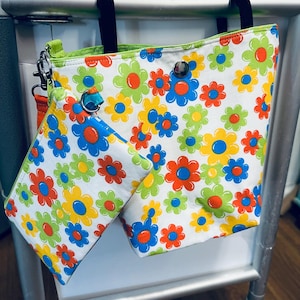 May include: A white tote bag and a matching smaller pouch, both featuring a repeating pattern of stylized flowers in bright shades of lime green, yellow, orange, and blue. The tote has black fabric straps and a silver metal snap closure.