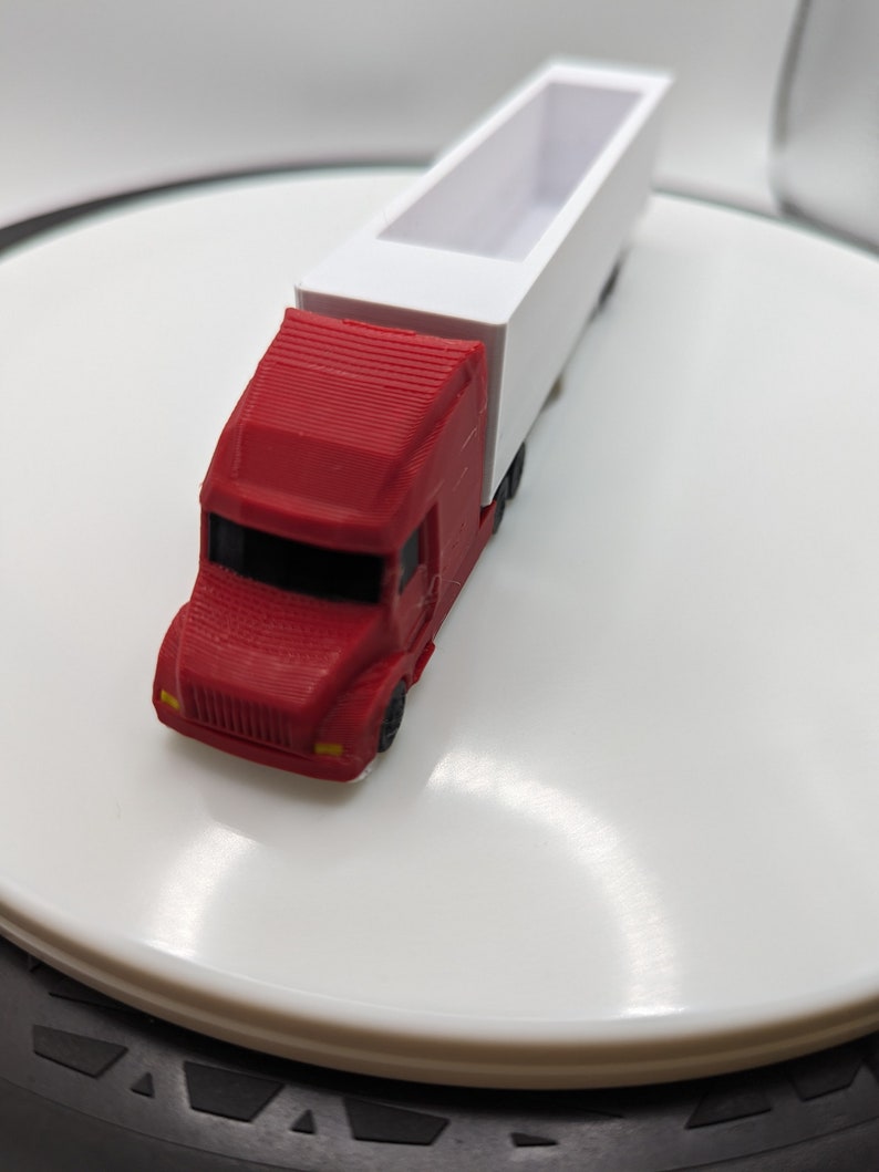 May include: A red toy semi-truck with a white trailer. The truck has a detailed cab and wheels. The trailer is empty.