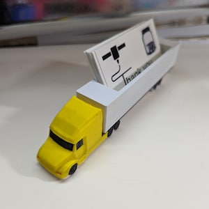 May include: A yellow toy semi-truck with a white trailer. The trailer is holding a stack of business cards with the text "Thank you" printed on them.
