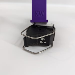 May include: A black car switch with a silver metal frame and a white symbol of a car with an arrow pointing to the right. The switch is labeled "OFF" and has a purple label with the letters "KW" on top.
