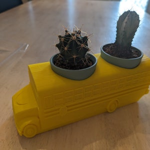 Puede incluir: Un macetero con forma de autobús escolar amarillo con dos pequeñas plantas de cactus en macetas grises. El autobús tiene un diseño detallado con ventanas y ruedas.