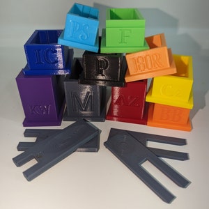 May include: A collection of colorful, 3D-printed boxes and supports. Each box is a different color, including blue, green, orange, and purple, and features embossed letters. The supports are gray.
