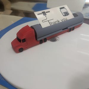 May include: A red and gray toy semi-truck with a trailer. The truck has a black windshield and black wheels. The trailer has a gray tank with a black cap. The truck is on a white surface. The truck has a small sign on the back that says "Thank you".