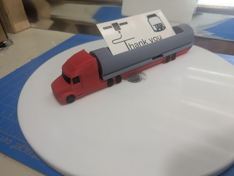 May include: A red and gray toy semi-truck with a trailer. The truck has a black windshield and black wheels. The trailer has a gray tank with a black cap. The truck is on a white surface. The truck has a small sign on the back that says "Thank you".