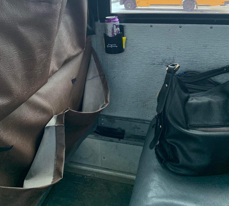 Op de afbeelding: Een zwarte plastic bekerhouder met het label "Mountain Cup Caddy" gemonteerd op een grijze wand, met daarin een paars-wit drankblikje. Links is een bruine, gestructureerde rugleuning met opbergvakken en rechts een zwarte handtas te zien.