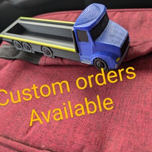 May include: A blue and gray toy truck with a flatbed. The truck is sitting on a red surface with the text "Custom orders available" in yellow.