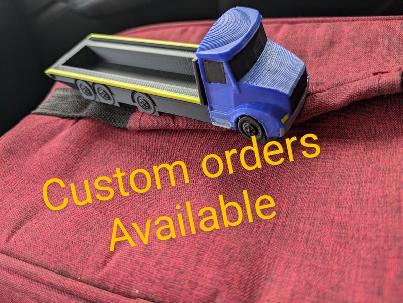 May include: A blue and gray toy truck with a flatbed. The truck is sitting on a red surface with the text "Custom orders available" in yellow.