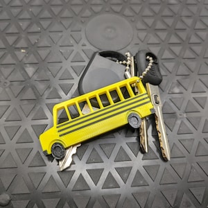 May include: A yellow school bus keychain with black stripes and wheels. The keychain is attached to a silver chain and a black key fob.