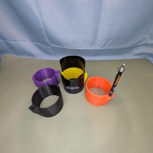 Op de afbeelding: Vier cilindervormige bekerhouders in paars, zwart, geel en oranje. De zwarte houder is voorzien van de tekst Magnetic Dip Caddy. Een oranje houder heeft een zwarte pen met een zilveren clip in een kleine zijbevestiging aan de rand gestoken.